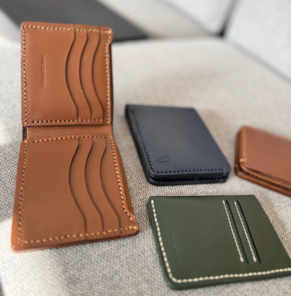 Brown leather bifold wallet, black wallet, and green card holder arranged on grey fabric.